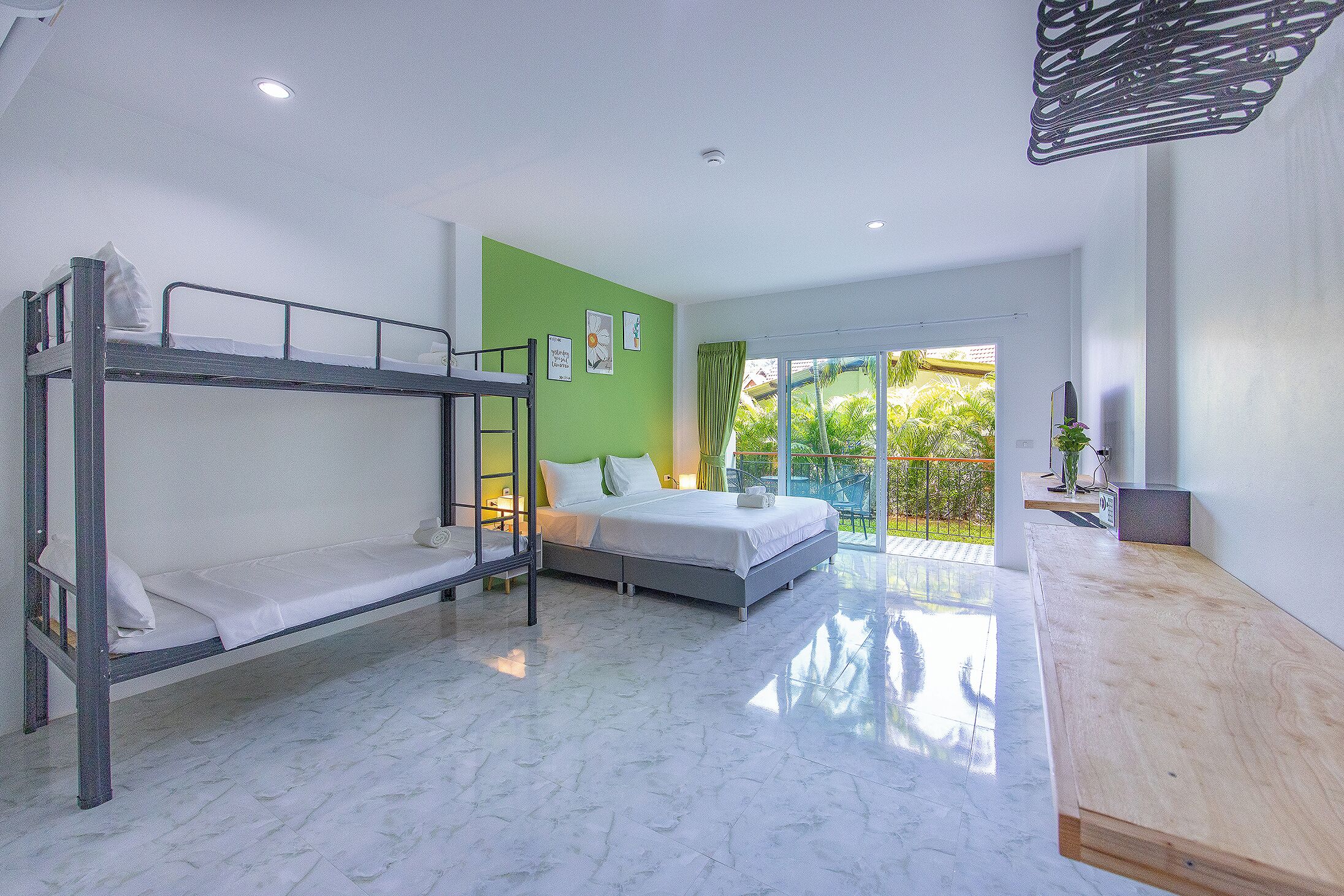 A modern bedroom with bunk beds, a large bed, a wooden desk, and a view of greenery through the glass door.