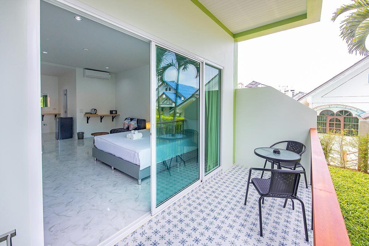 A modern hotel room with a large bed, a glass sliding door leading to a balcony with a table and chairs, and a view of the surrounding buildings.