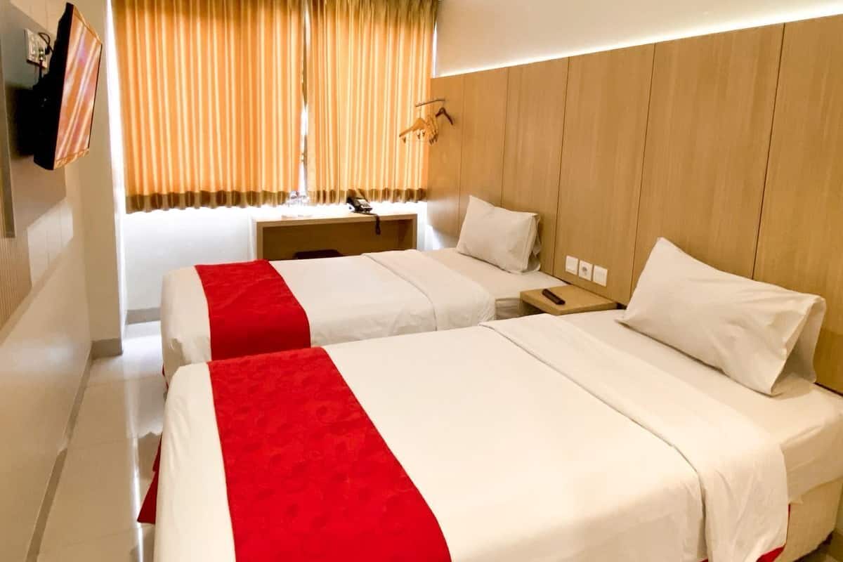 Ein Hotelzimmer mit zwei Betten, einem hölzernen Kopfteil, einem Schreibtisch und einem großen Fenster mit Vorhängen.