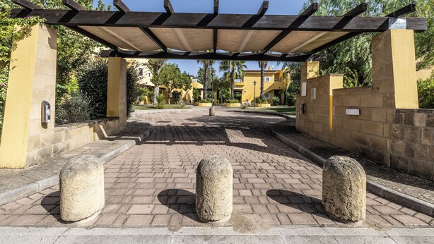 Een verharde oprijlaan met een pergola, stenen paaltjes en gele gebouwen op de achtergrond.