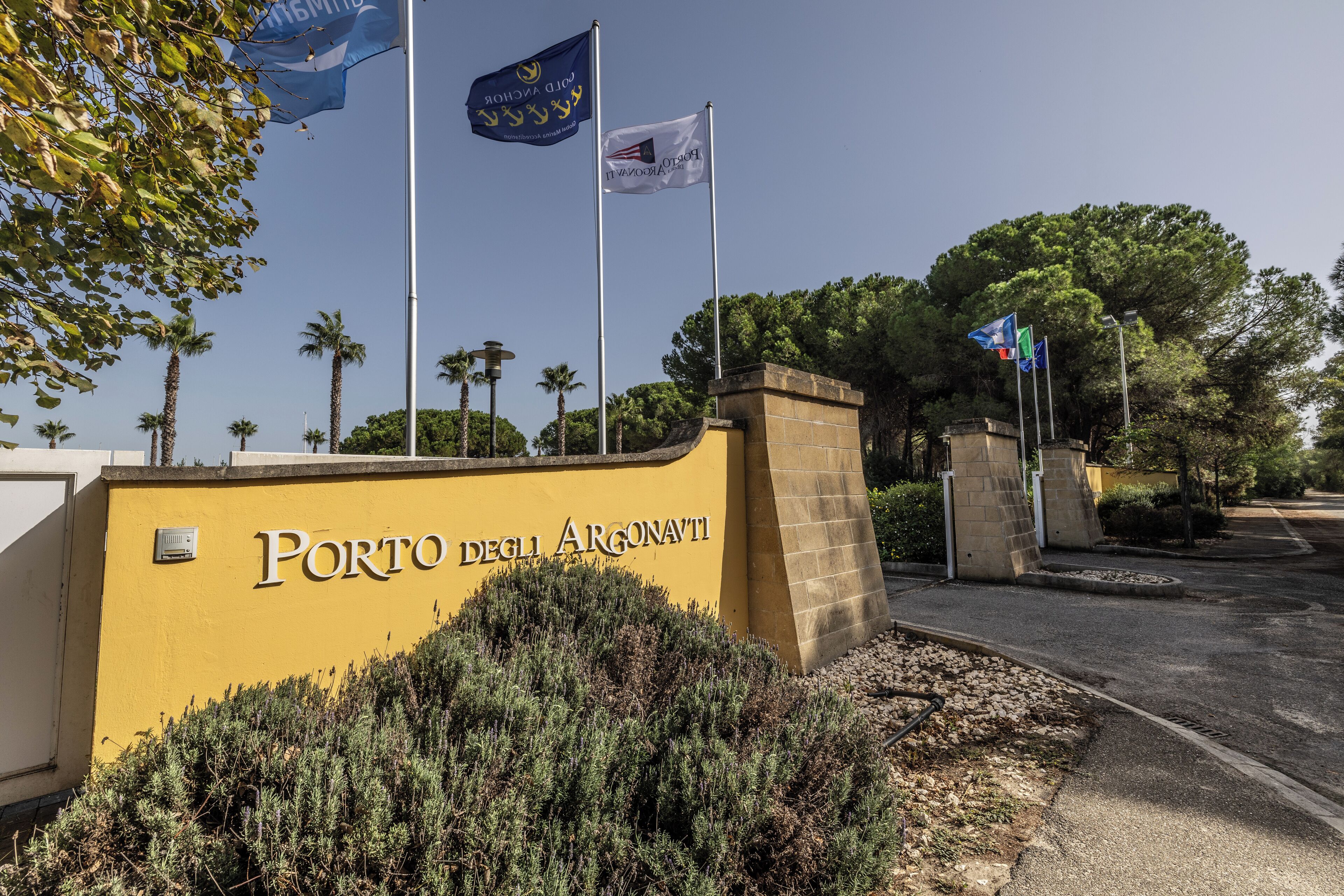 A yellow wall with the text "Porto degli Argonauti" and multiple flags, including the European Union flag.