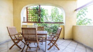 Een overdekt terras met een houten tafel en stoelen, een boogvenster en uitzicht op groen.