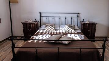 A neatly made bed with a patterned bedspread and pillows, flanked by two wooden nightstands with lamps, in a room with wooden flooring and a plain wall.