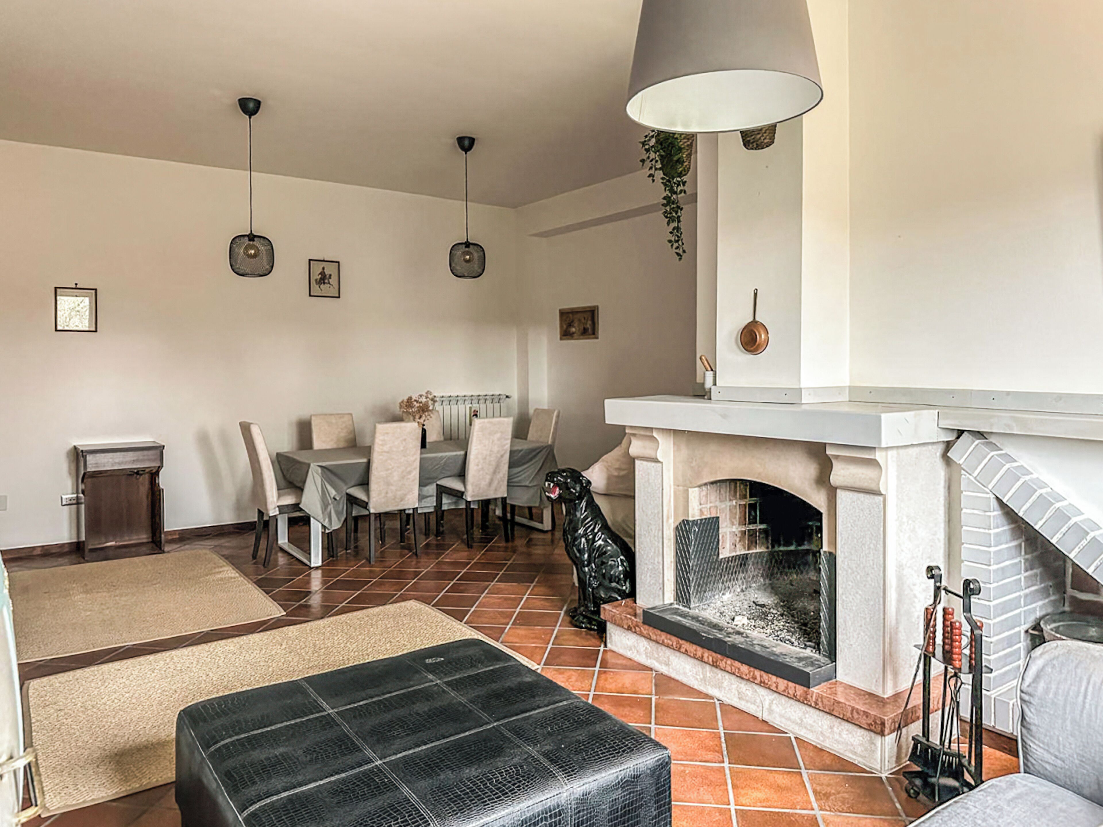Una sala de estar con chimenea, una mesa de comedor con sillas, un perro negro y una lámpara colgante.