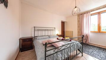 A bedroom with a metal bed frame, a bedside table, a nightstand, a window with curtains, and a patterned rug.