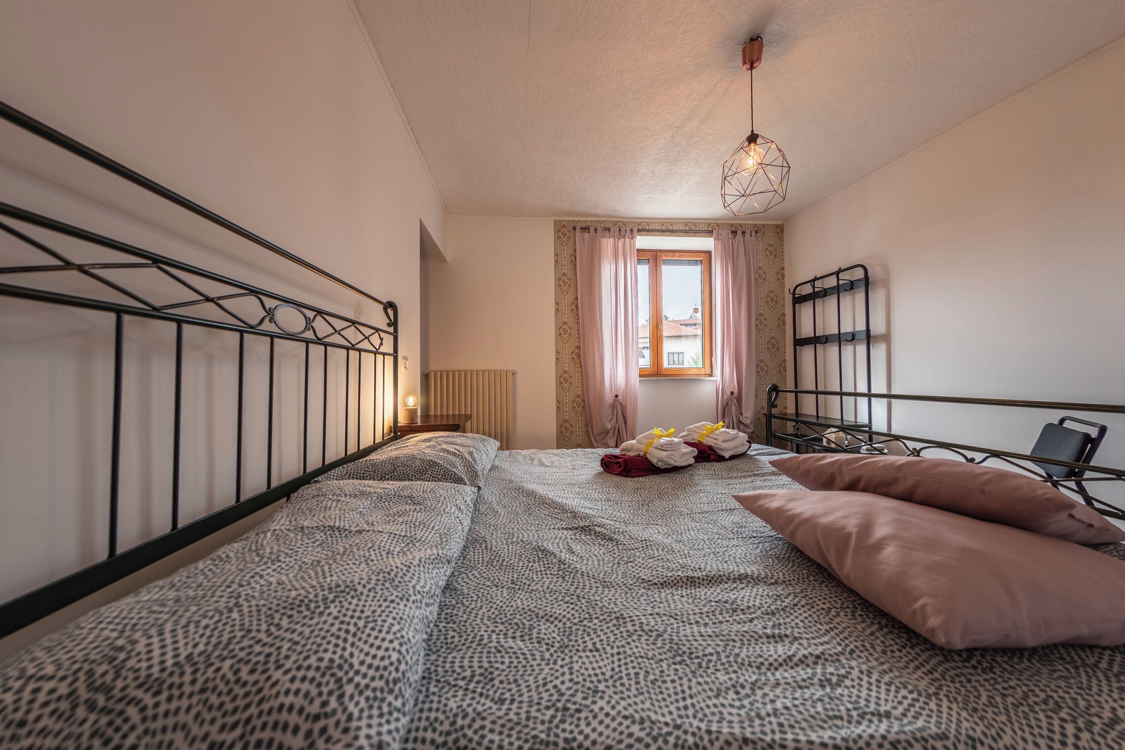 Un dormitorio con cama, mesita de noche, ventana con cortinas y lámpara colgante.