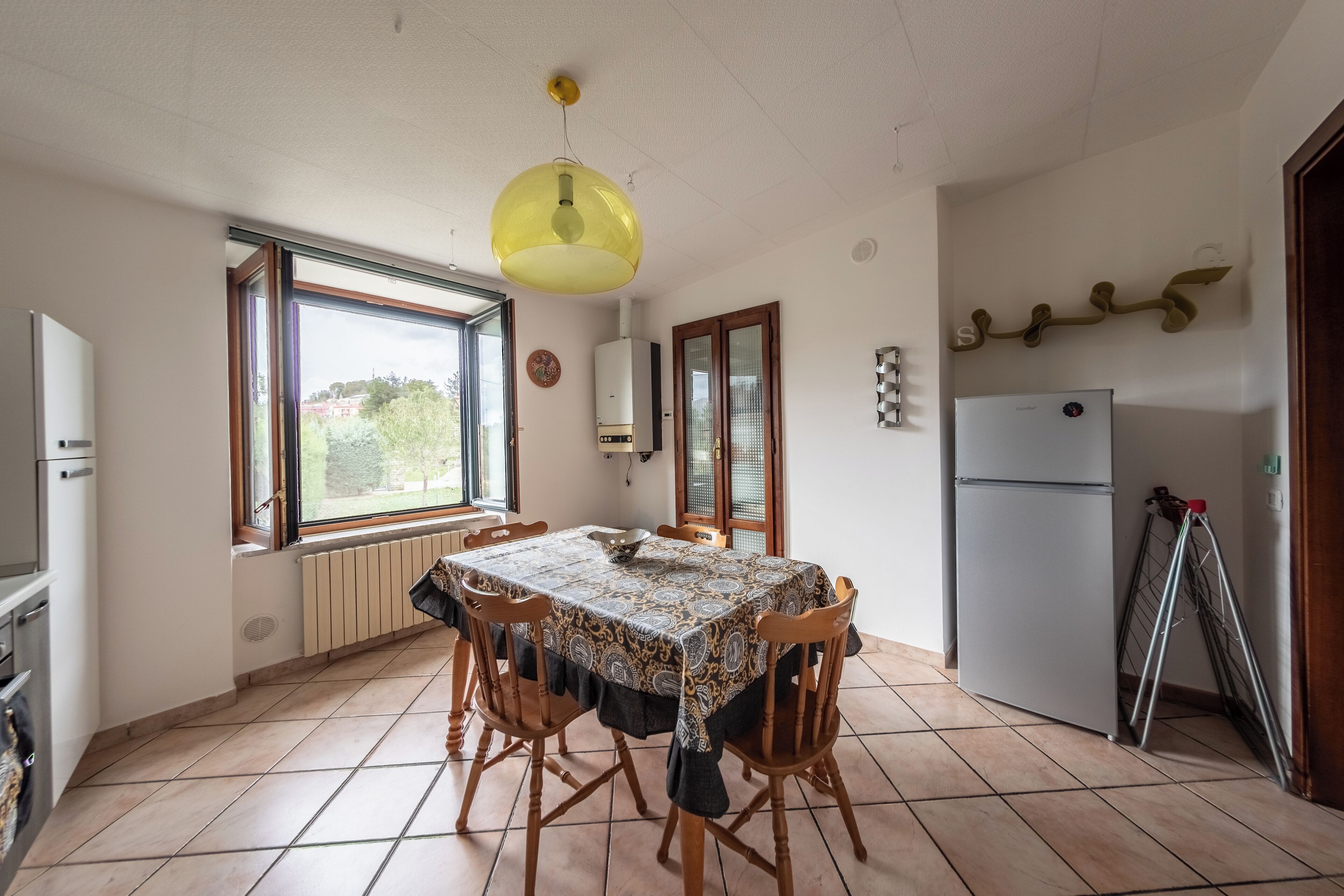A kitchen with a dining table, chairs, a hanging light fixture, a refrigerator, and a window with a view of greenery.