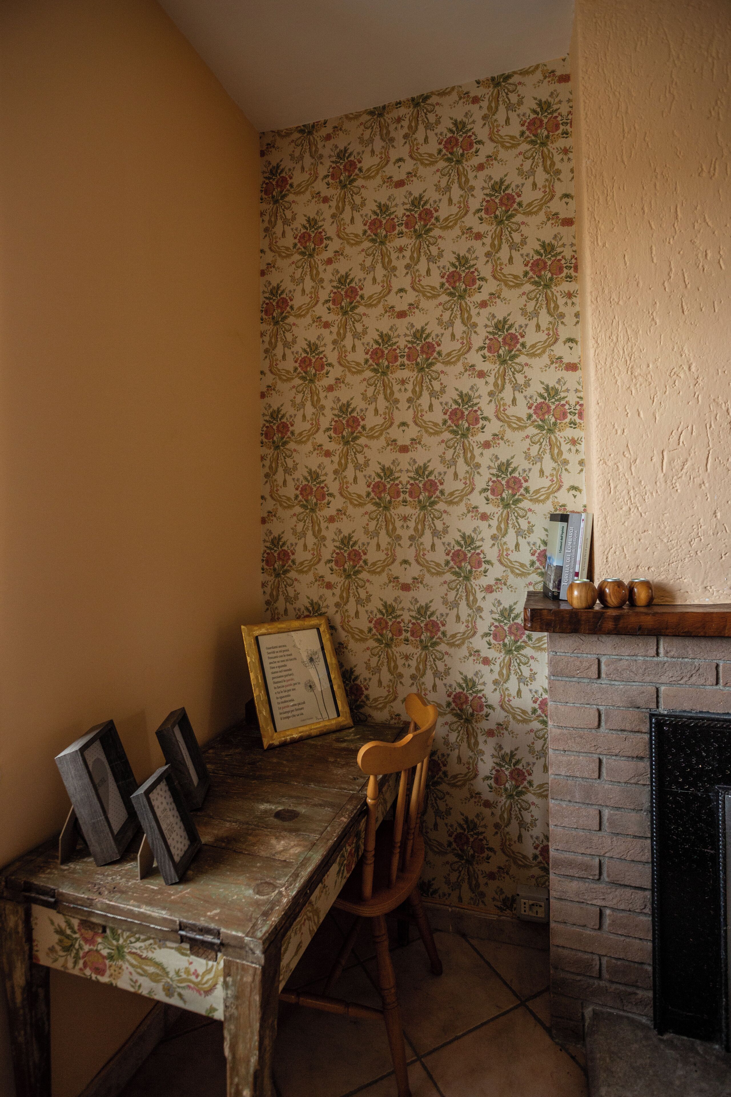 Una habitación de esquina con papel tapiz floral, una mesa de madera con marcos de cuadros, una silla y una chimenea.
