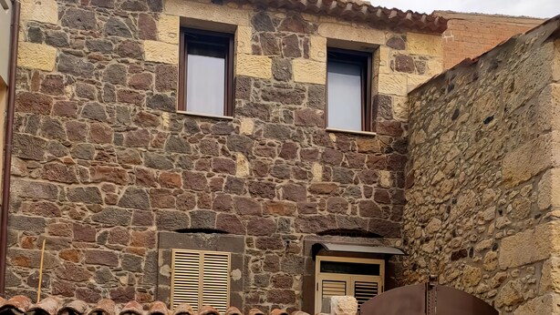 A stone building with a tiled roof, a wooden door with a sun design, and a small window. - Favisi Rooms (Sagama)