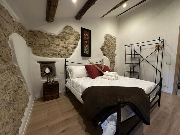 A bedroom with a stone wall, a bed with white bedding and a brown throw, a nightstand with a mirror, and a metal canopy bed frame.