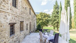 A stone building with a patio table set for dining, surrounded by greenery and trees. - Il Casale Di John (Umbertide)