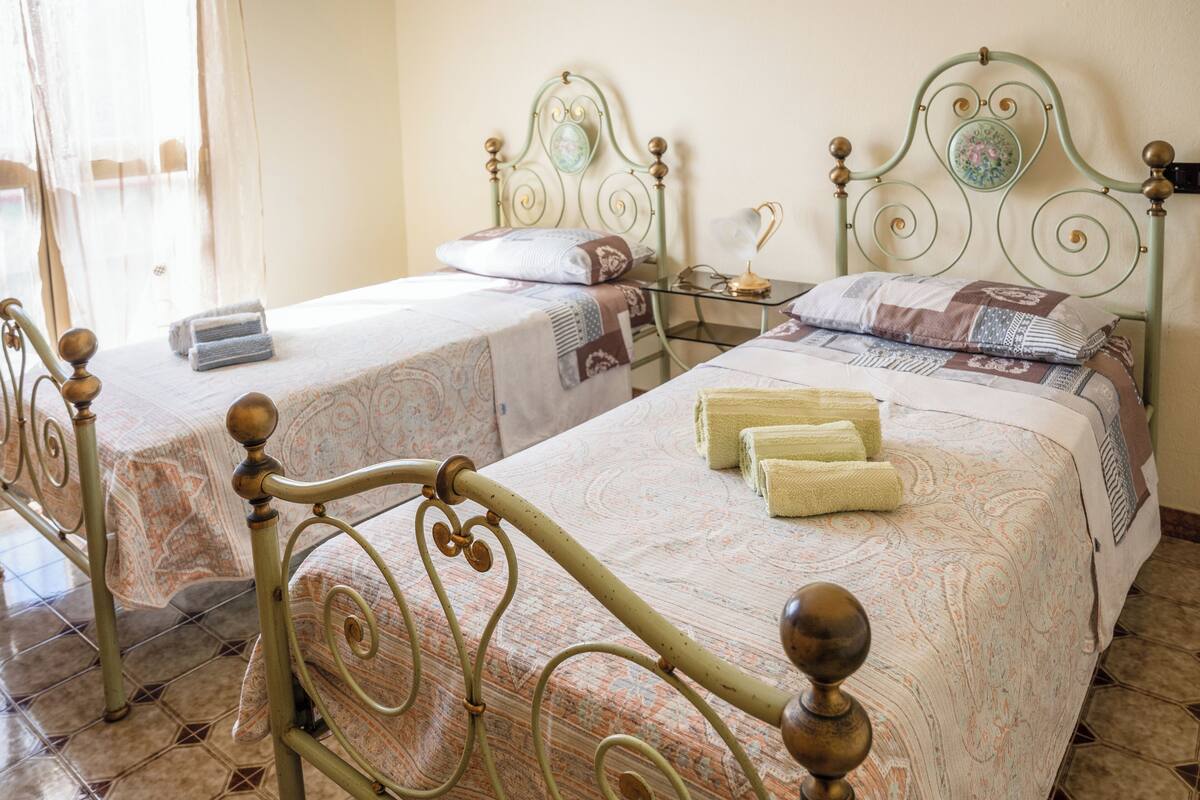 A room with two single beds, each with a distinct headboard design. There is a small table between the beds with a lamp on it.