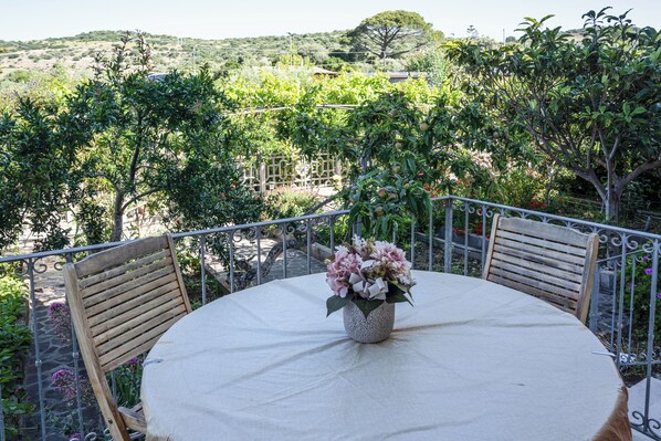 Een ronde tafel met een wit tafelkleed, twee houten stoelen en een bloevas op een balkon met metalen hekwerk en uitzicht op bomen en heuvels.