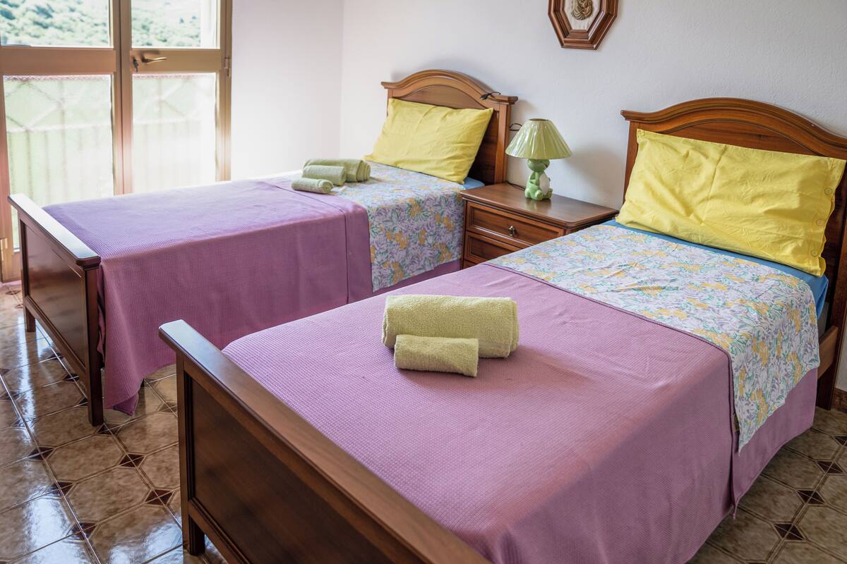 Two single beds with wooden headboards, each with a purple bedspread and a yellow pillow. There is a wooden nightstand with a green lamp between the beds.