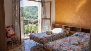 A bedroom with two beds, a chair, and a view of a green landscape through an open door. - Le Case nel Vitigno (Modolo)