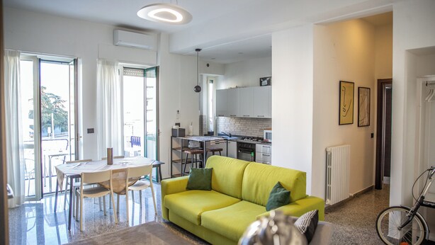 A modern living room with a yellow sofa, dining table, and chairs. There is a kitchen area with white cabinets and a bicycle in the corner. - Billotti Central Loft - Cosenza (Cosenza)