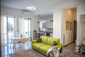 Ein modernes Wohnzimmer mit einem gelben Sofa, einem Esstisch und Stühlen. Es gibt eine Küchenzeile mit weißen Schränken und ein Fahrrad in der Ecke.