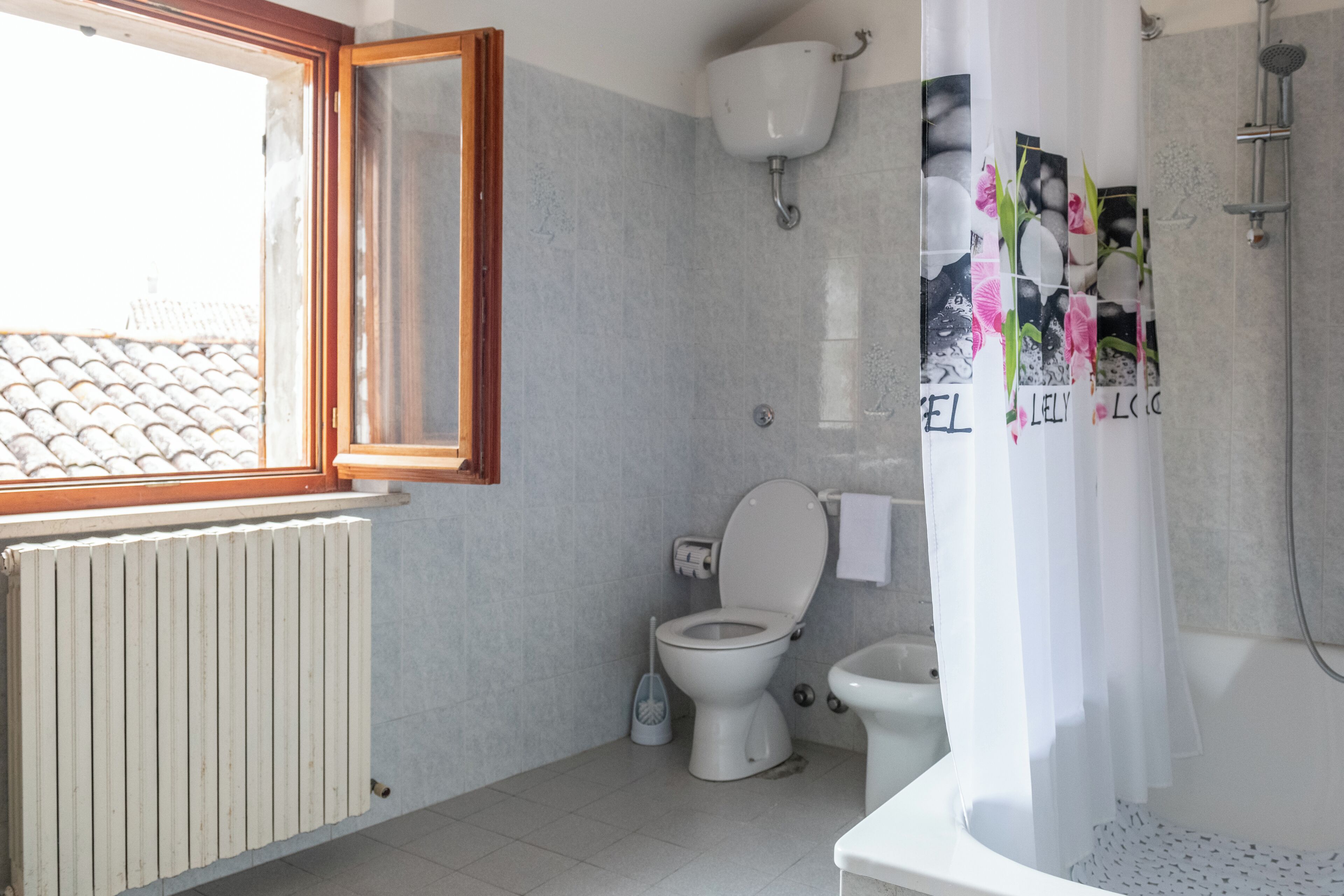 Een badkamer met toilet, bidet en douche. Er is een raam met houten kader en een radiator.
