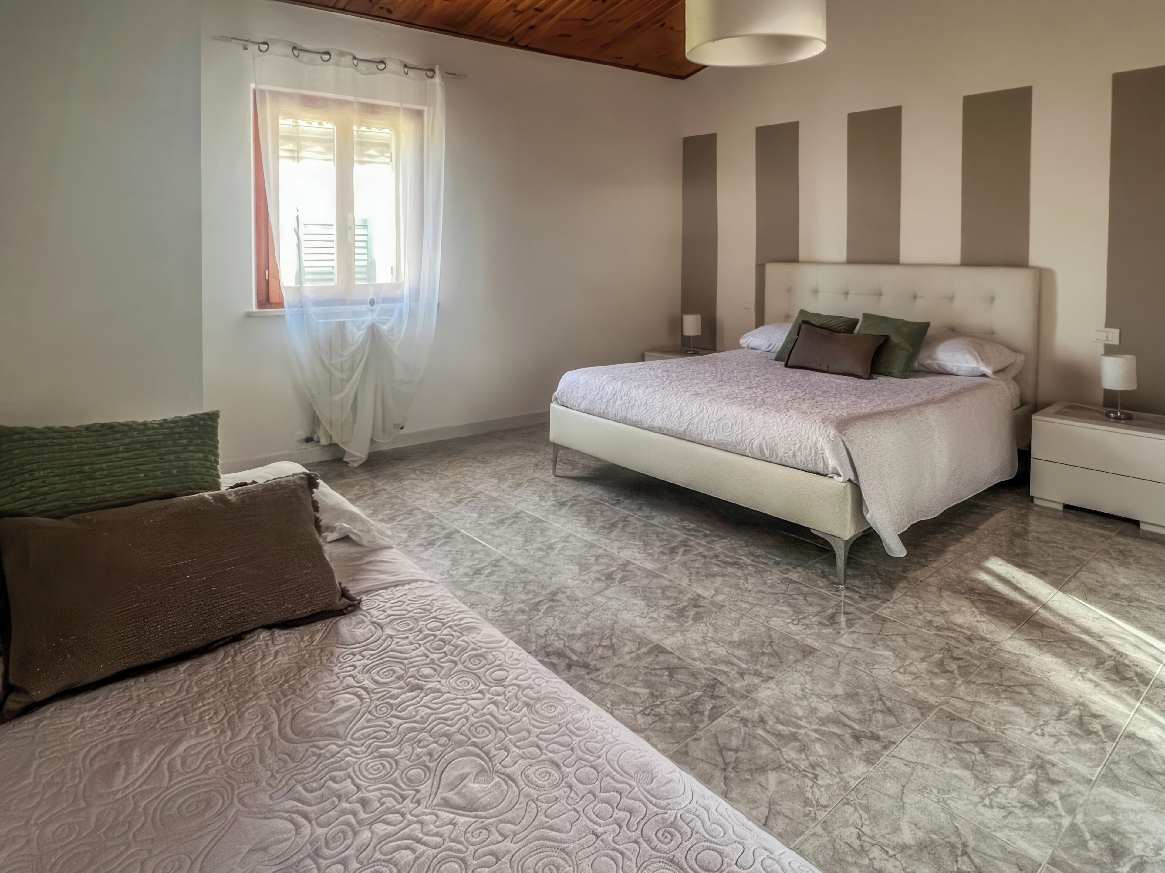 Un dormitorio con una cama, una mesita de noche, una lámpara, una ventana con cortinas y techo de madera.