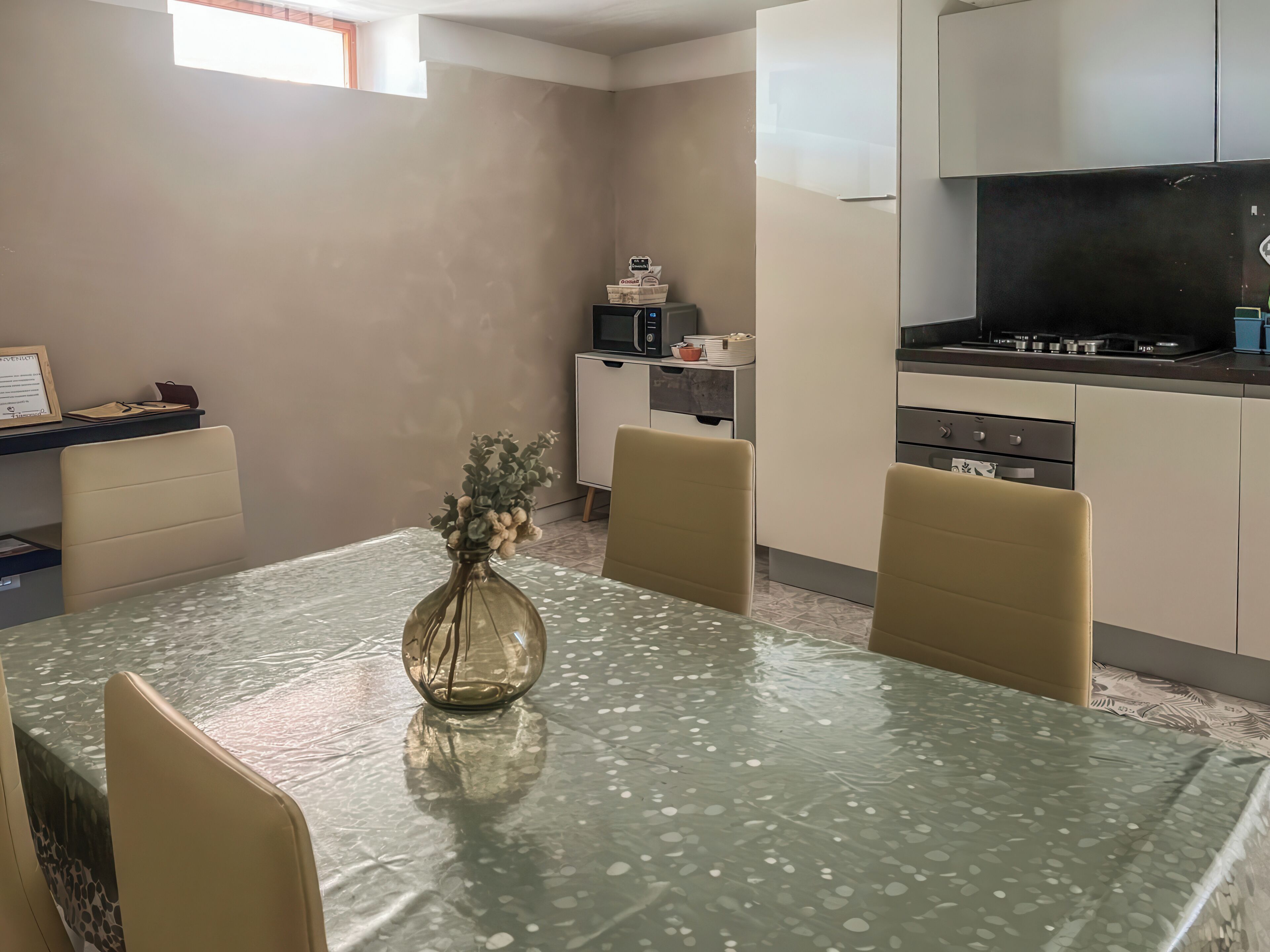 A dining area with a table, chairs, a vase with flowers, a kitchen area with appliances, and a cabinet.