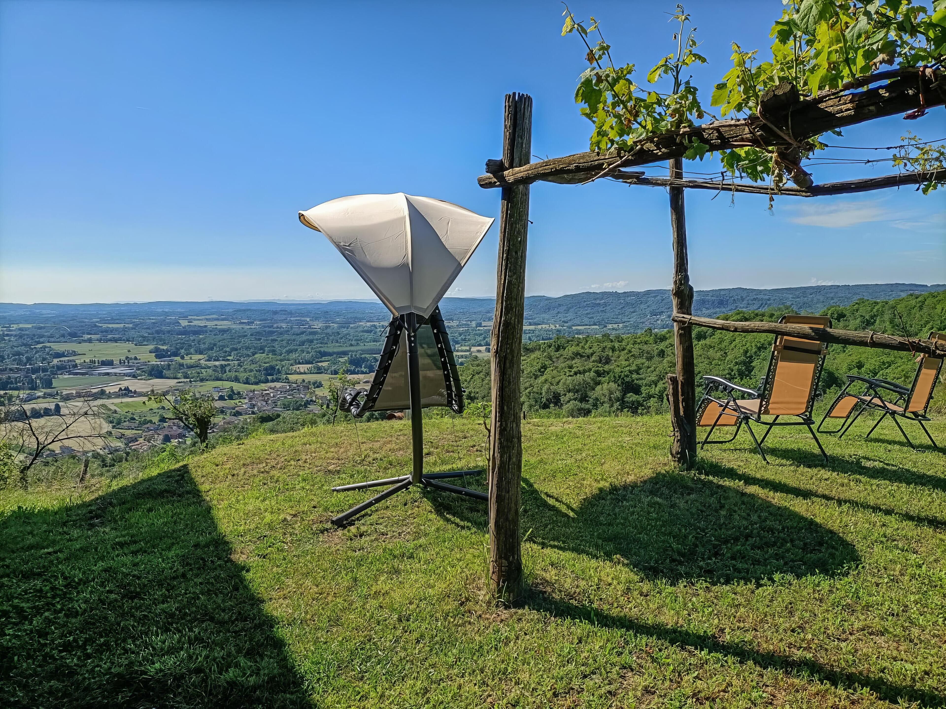 Un'antenna parabolica su un treppiede, un pergolato in legno e una fila di sedie su una collina erbosa con vista su una valle e colline lontane.