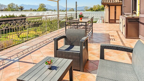 Eine Terrasse mit Korbmöbeln, einem Tisch mit Topfpflanze und Blick auf ein Gewächshaus und Berge.