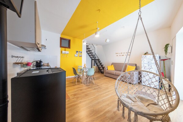 Una cocina moderna con un refrigerador negro, una zona de comedor con una mesa y sillas, una silla colgante y una escalera que lleva a una sala de estar con un sofá y un muro decorativo.
