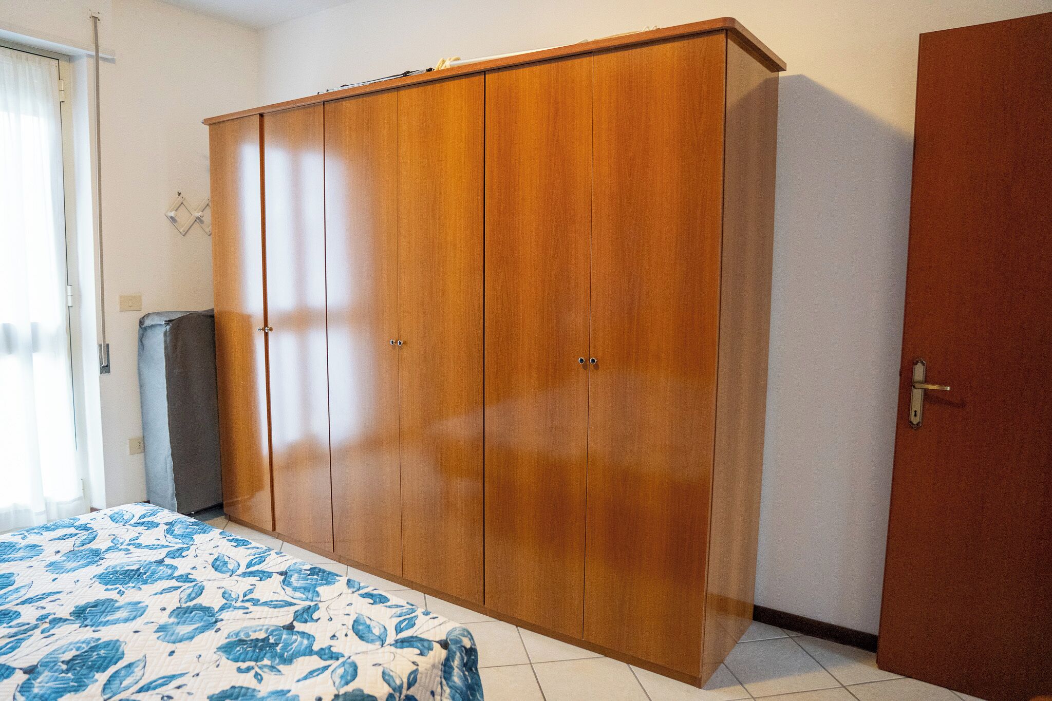 Un dormitorio con un armario de madera grande, una cama con colcha estampada y una puerta a la derecha.