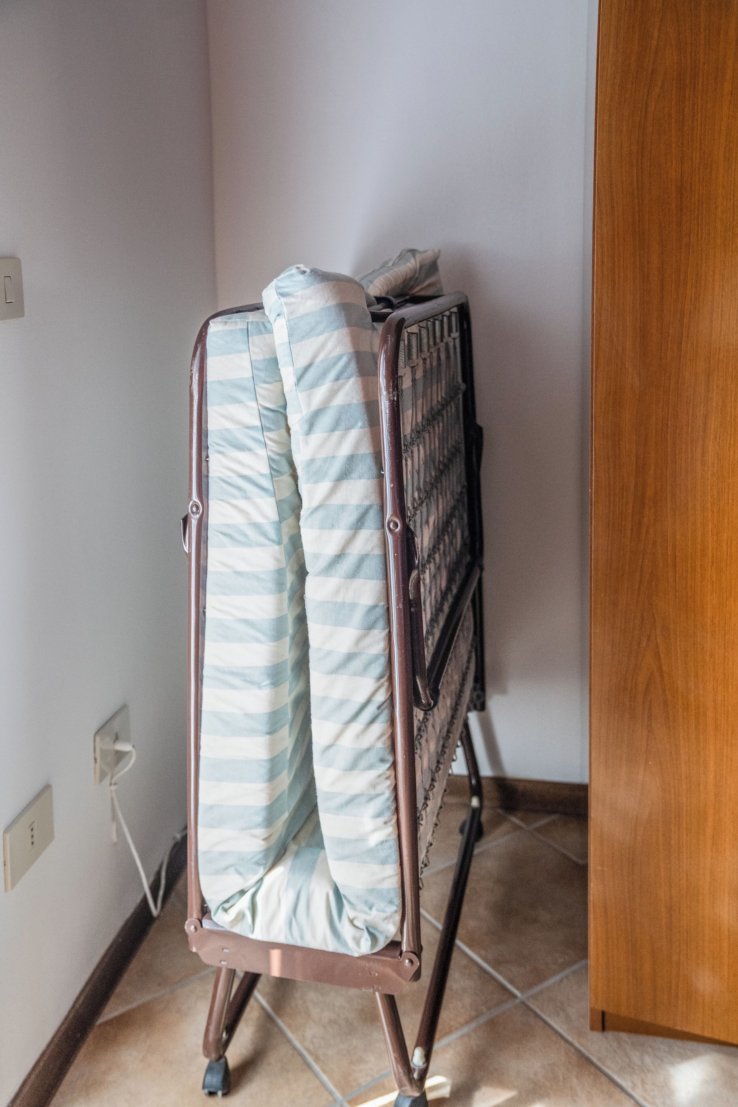 Esquina de una habitación con un estante metálico que sostiene un colchón y almohadas, una puerta de madera y un suelo de baldosas.