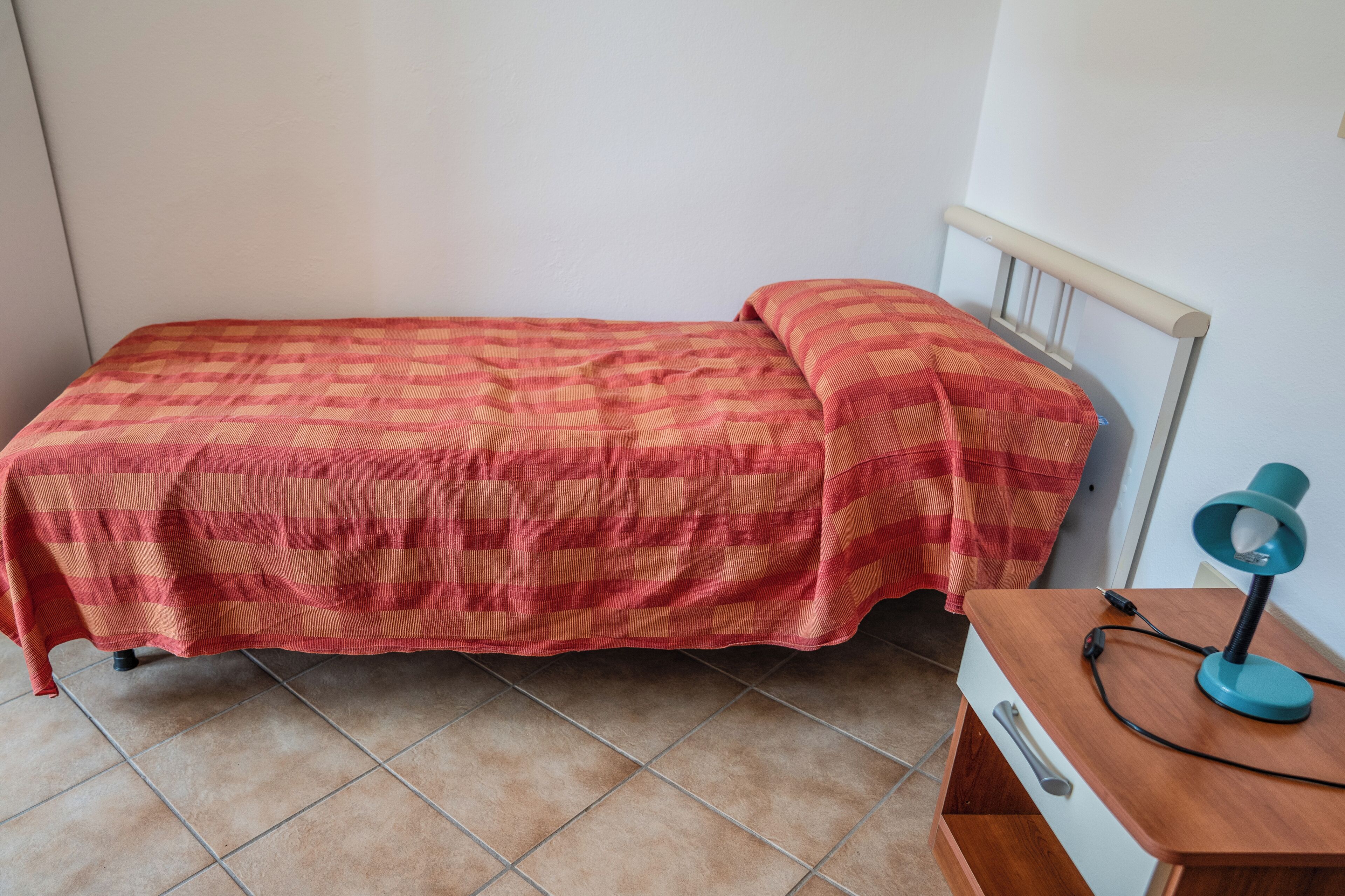 Un lit simple avec une couvre-lit à carreaux, une table de chevet avec une lampe et une petite table en bois.