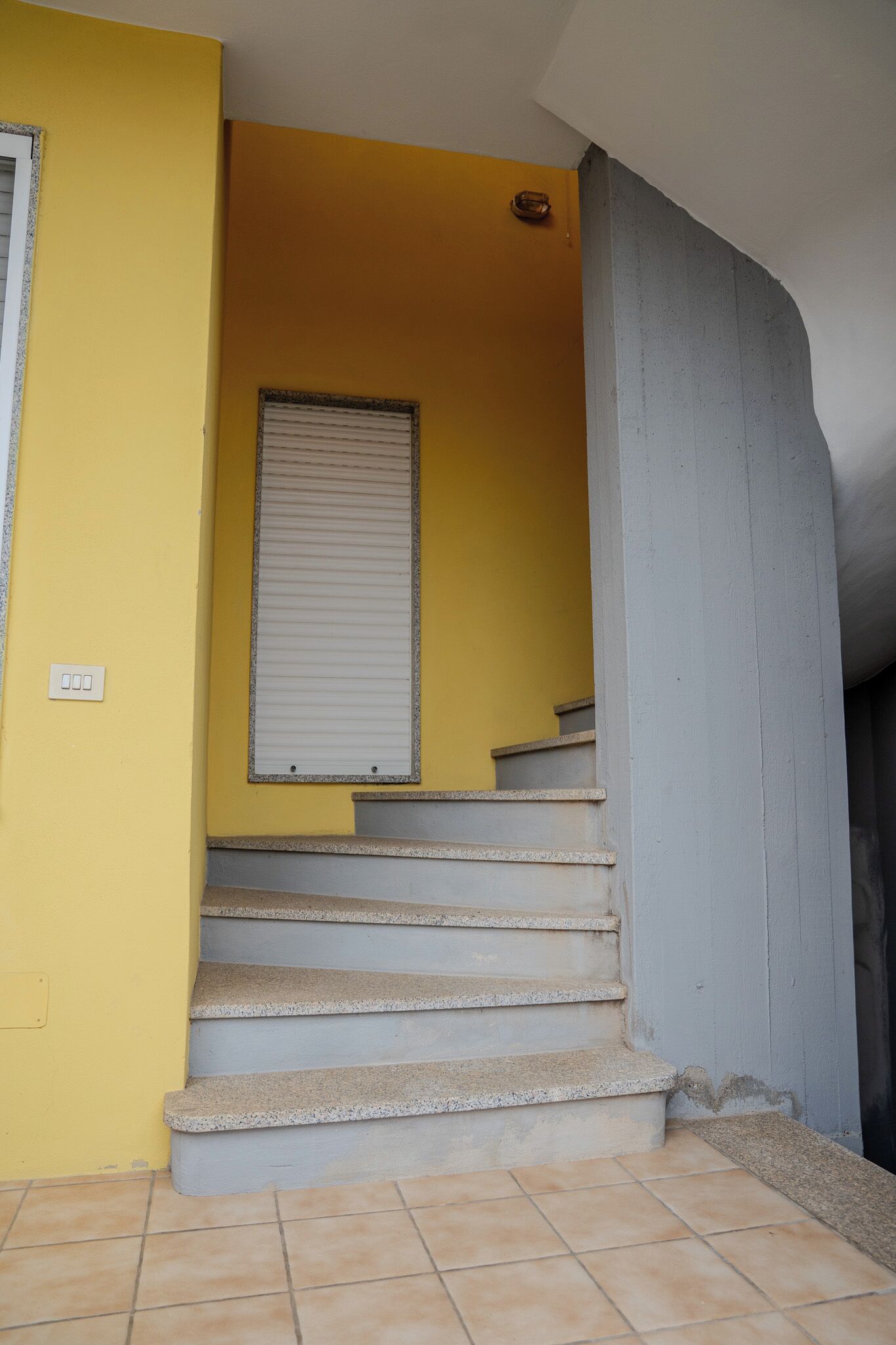 A staircase with a yellow wall and a window with a closed shutter.