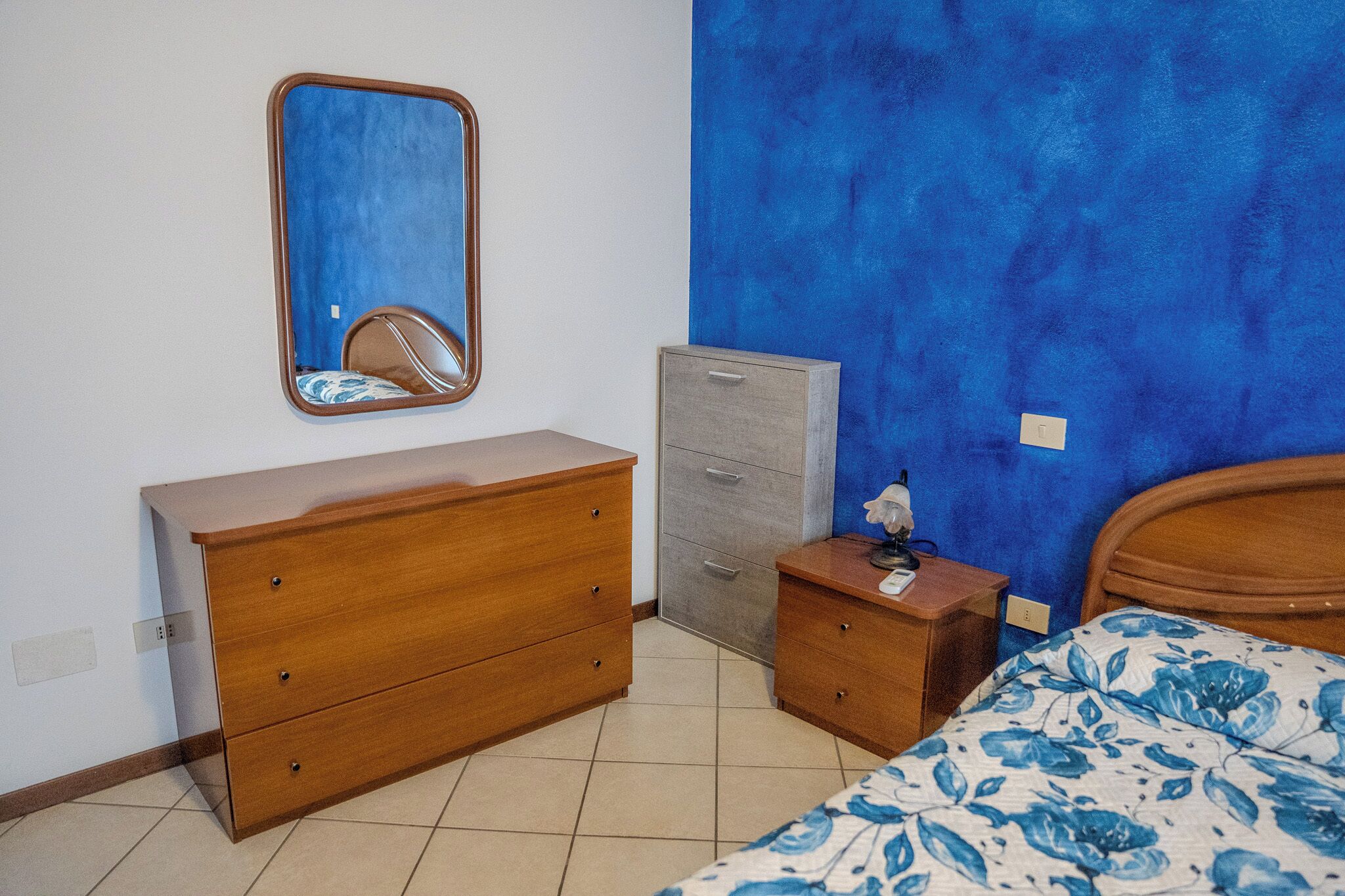 Un dormitorio con un aparador de madera, una mesita de noche pequeña, una mesita auxiliar, una cama con colcha estampada y un espejo, además de una pared azul decorativa.
