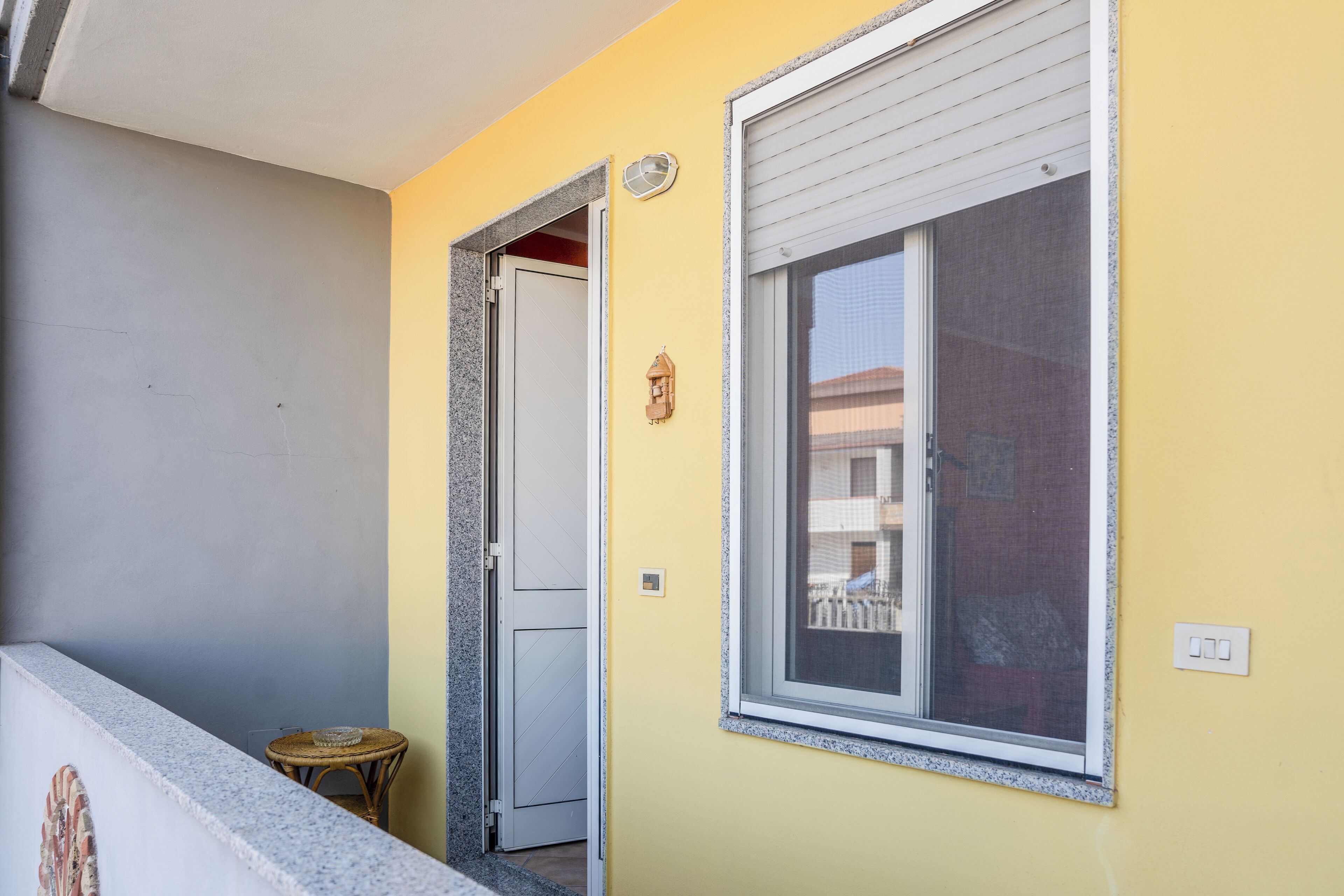 Ein Balkon mit einer weißen Tür, einem kleinen Korbstuhl und einem Fenster mit Fliegengitter.