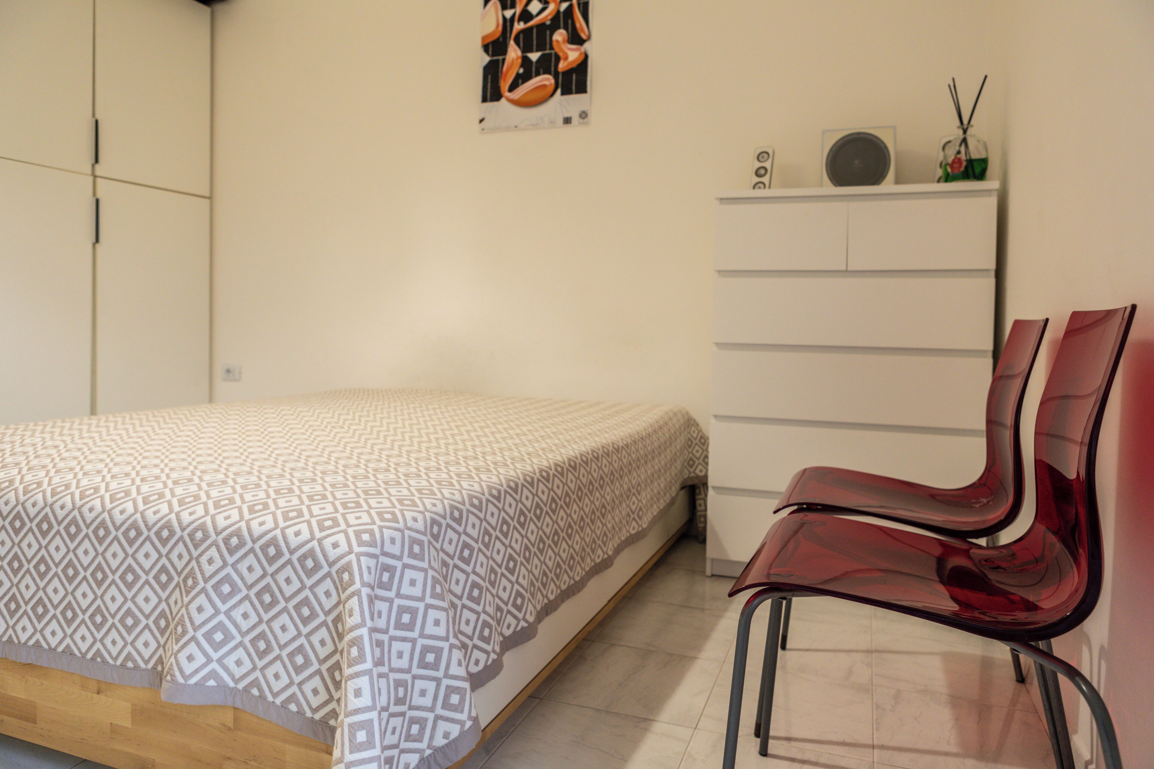 Une chambre à coucher comprenant un lit, un fauteuil rouge, une commode et une étagère murale avec un haut-parleur et un objet décoratif.