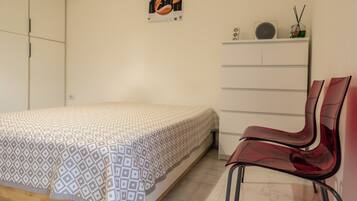 Une chambre à coucher comprenant un lit, un fauteuil rouge, une commode et une étagère murale avec un haut-parleur et un objet décoratif.