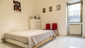 A bedroom with a bed, a dresser, two red chairs, and a window with a view of buildings. - Casa Frida (Roccagloriosa)