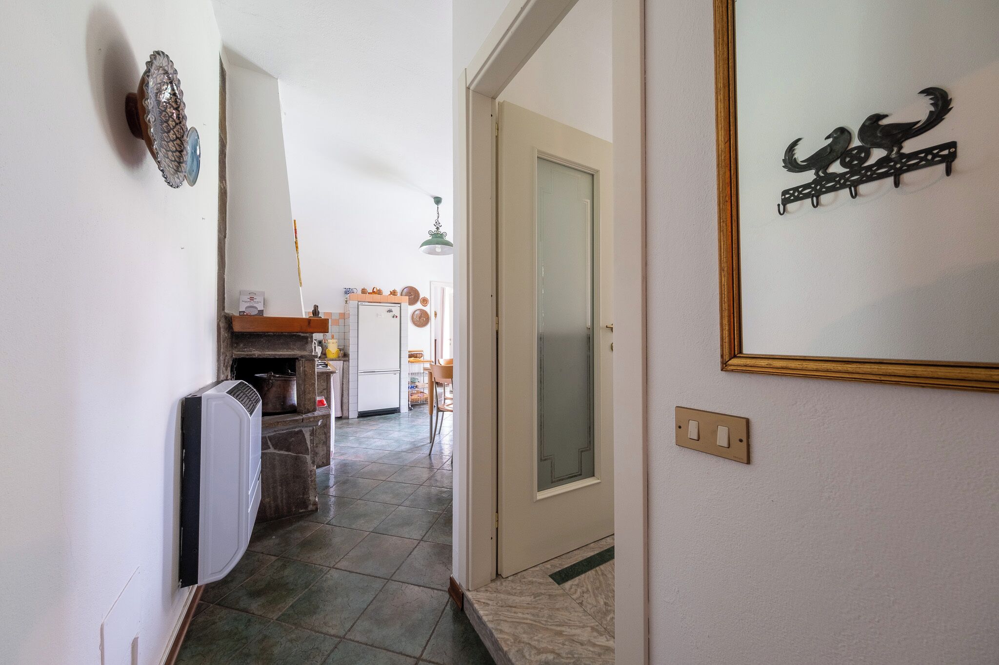 A narrow hallway with a kitchen area in the background, a door with a decorative metal piece, and a light switch on the wall.