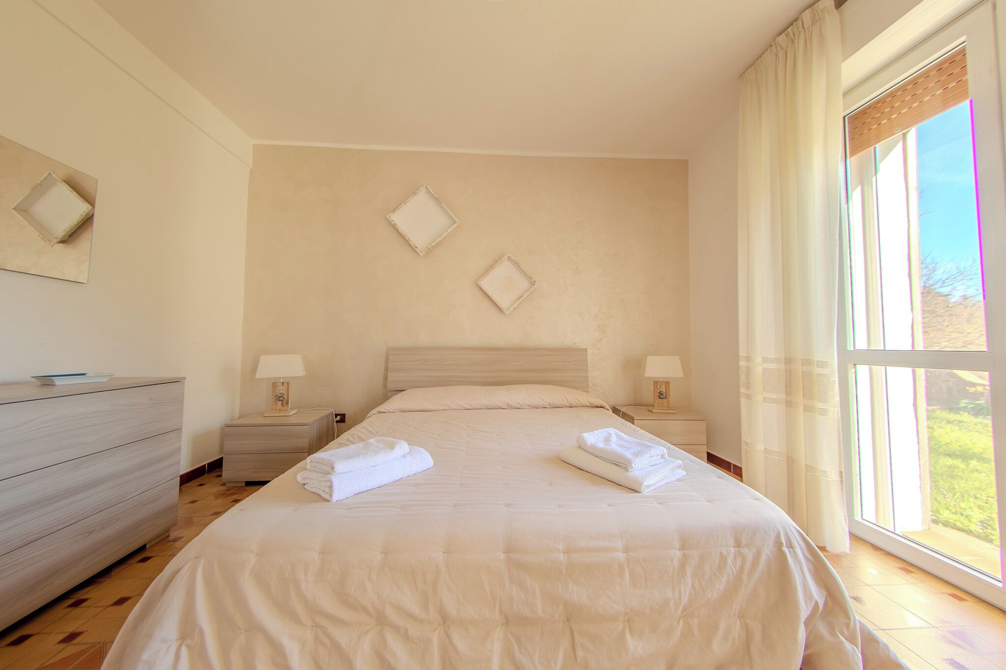 A neatly arranged bedroom with a large bed, two bedside tables with lamps, a dresser, and a window with curtains.