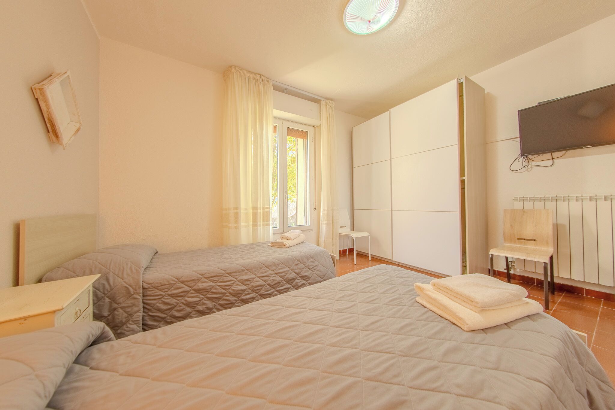 A bedroom with two beds, a desk, a chair, a television, and a window with curtains.