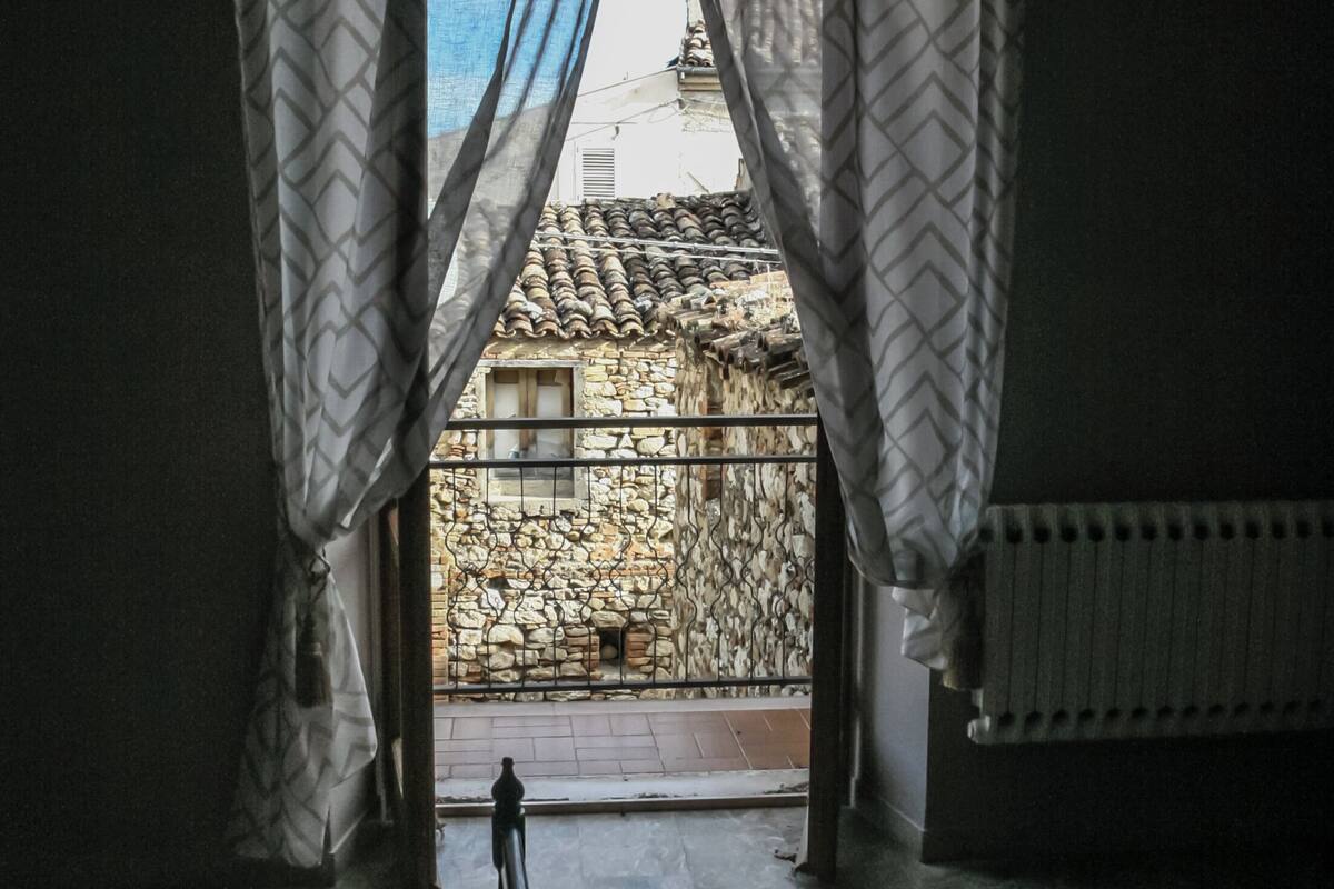A room with a bed, a window with patterned curtains, a view of a stone building outside, and a radiator on the wall.