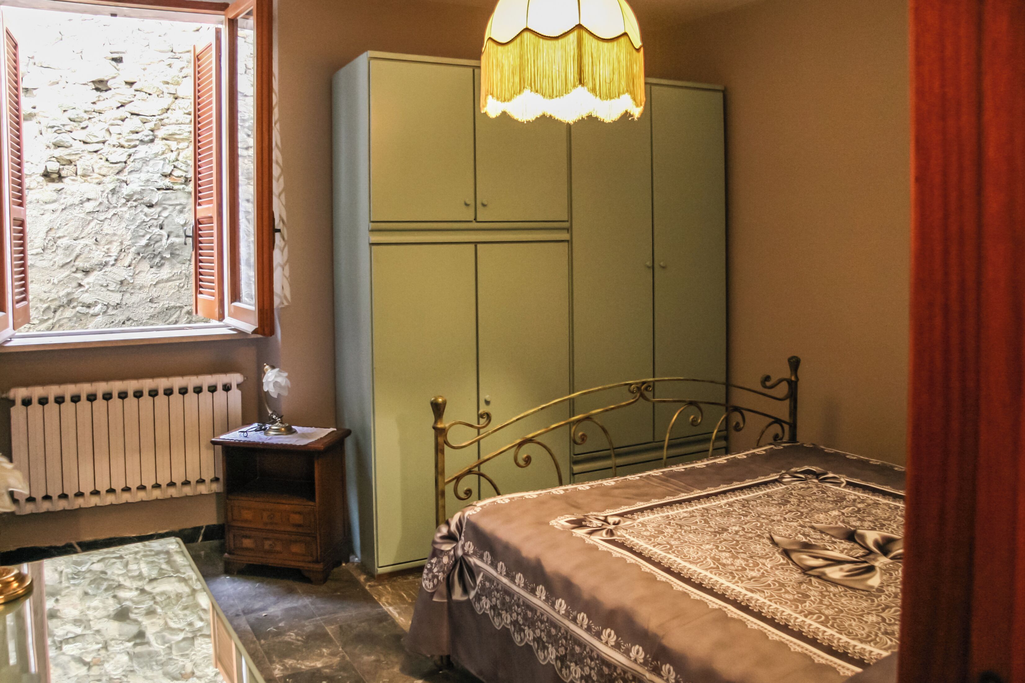 A bedroom with a bed, a wardrobe, a radiator, a small table with a lamp, and a window with shutters.