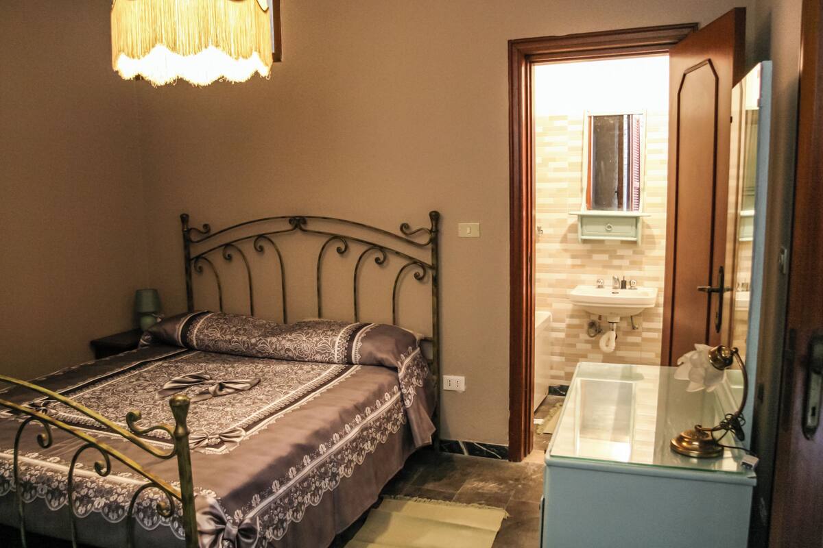 A bedroom with a bed, bedside table, lamp, and a bathroom with a sink and mirror.