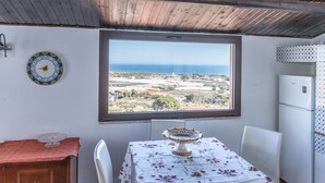 Ein Zimmer mit Tisch und Stühlen, einem Fenster mit Meerblick, einem Kühlschrank und einer Wanduhr.