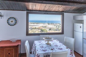 Una habitación con una mesa, sillas, una ventana con vista al mar, un refrigerador y un reloj en la pared.