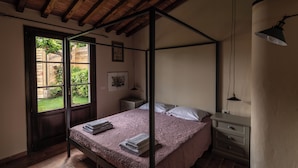 Een hemelbed met een roze sprei en witte kussens, een nachtkastje met een lamp, en een raam met uitzicht op groen.