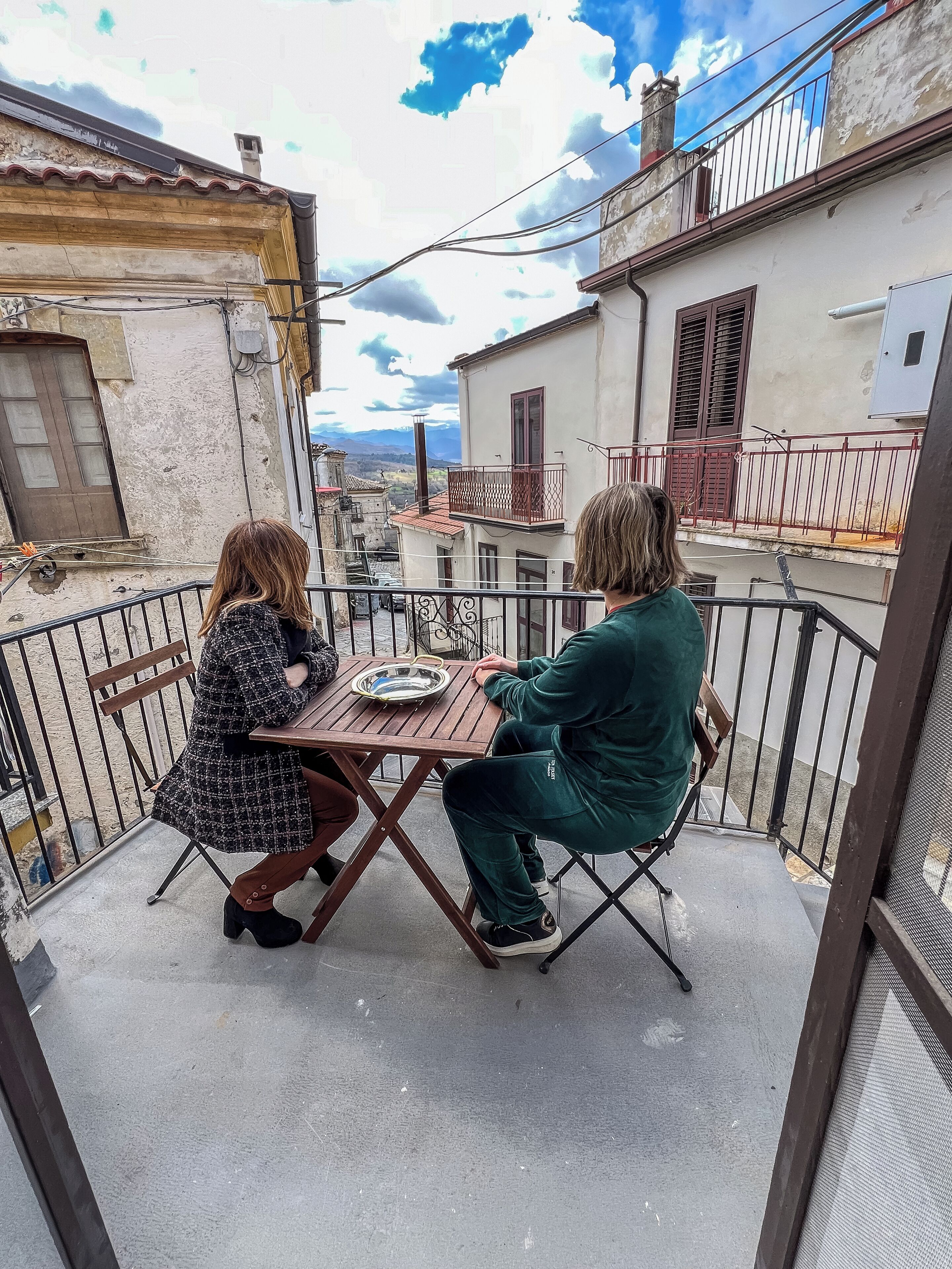 Two people seated at a table on a balcony with a view of buildings and mountains in the background.