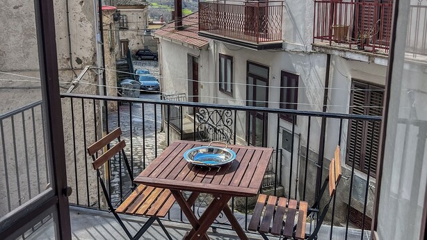 Un balcón con una mesa y sillas de madera, con vista a una calle con edificios y cielo despejado.