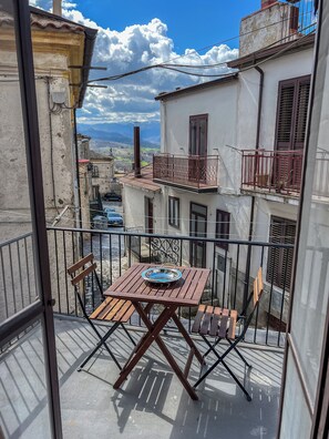 A balcony with a wooden table and chairs, overlooking a street with buildings and a clear sky. - Casetta San Giorgio Lucano (San Giorgio Lucano)