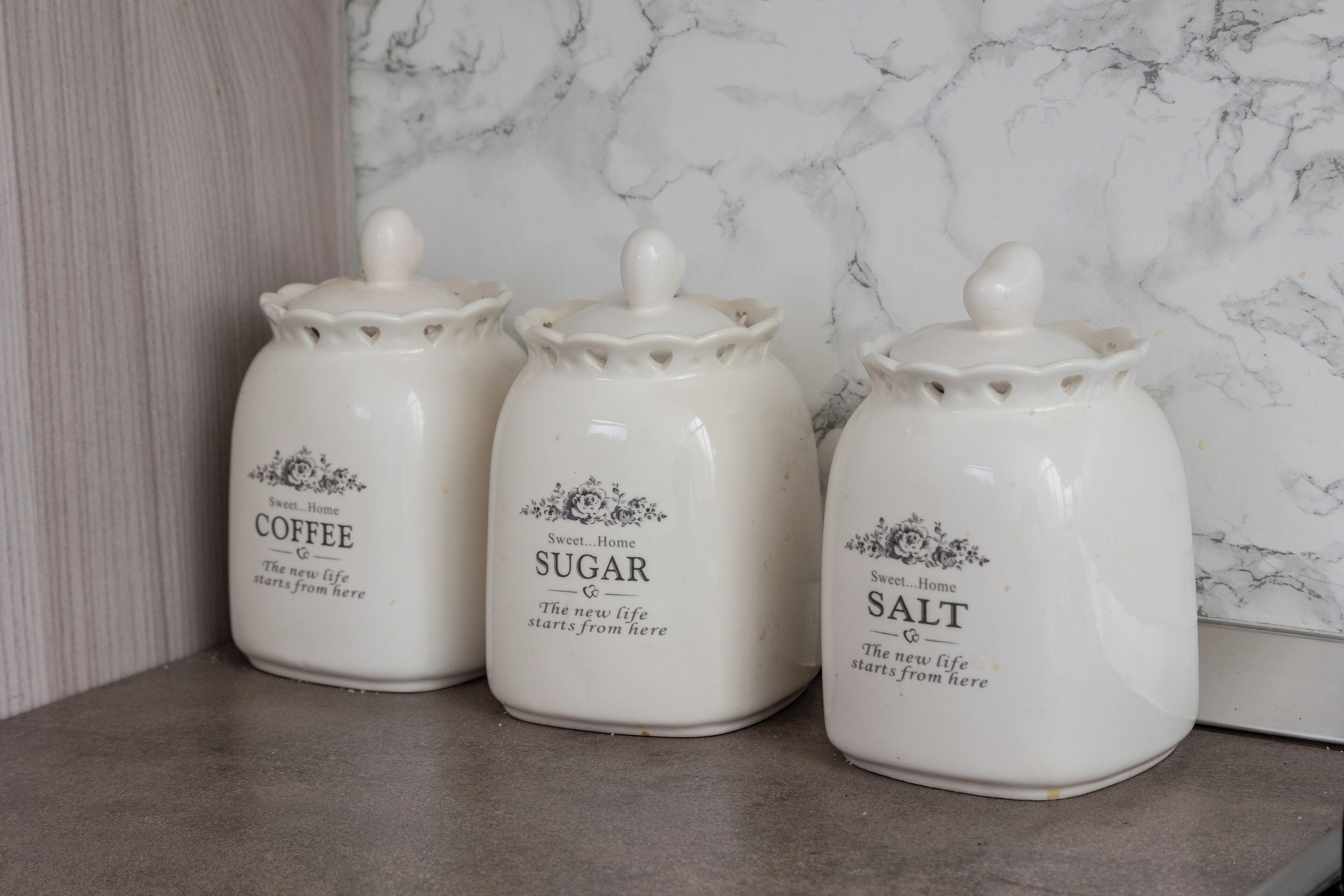 Tres recipientes de cerámica blanca con tapas, etiquetados 'Coffee', 'Sugar' y 'Salt', sobre una encimera de cocina con salpicadero de mármol.