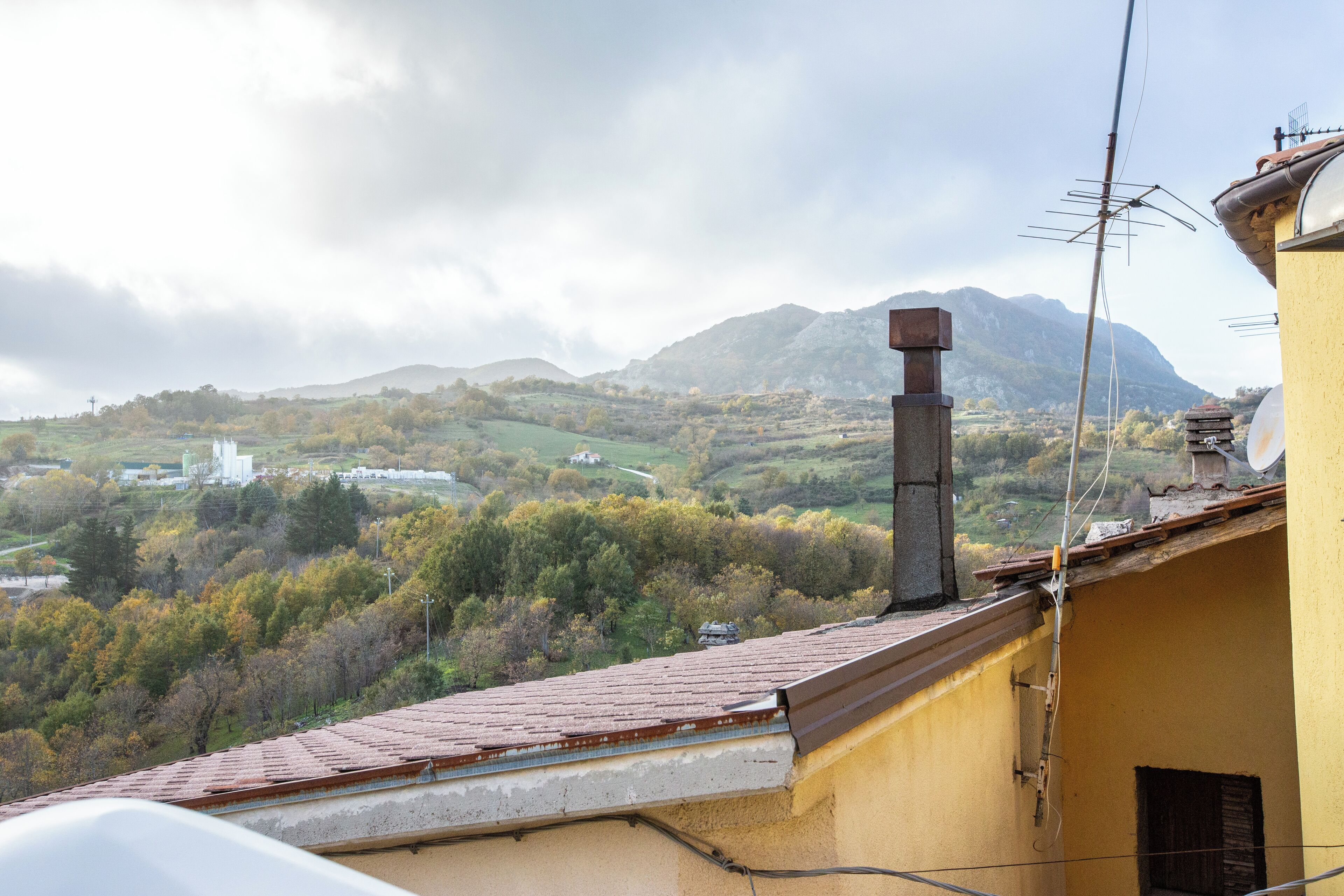 Ein Gebäude mit gelber Fassade, einem Schornstein und Blick auf eine hügelige Landschaft mit Bäumen und einer entfernten Struktur.